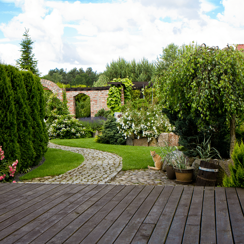 Jardin luxuriant avec terrasse en bois, chemin pavé sinueux, arches en briques fleuries, arbres verts et un chat roux près de plantes. Ciel nuageux.