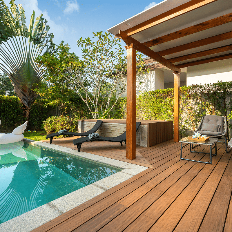 Terrasse en bois clair avec piscine turquoise, chaises longues, pergola ombragée et végétation tropicale luxuriante. Vue relaxante.