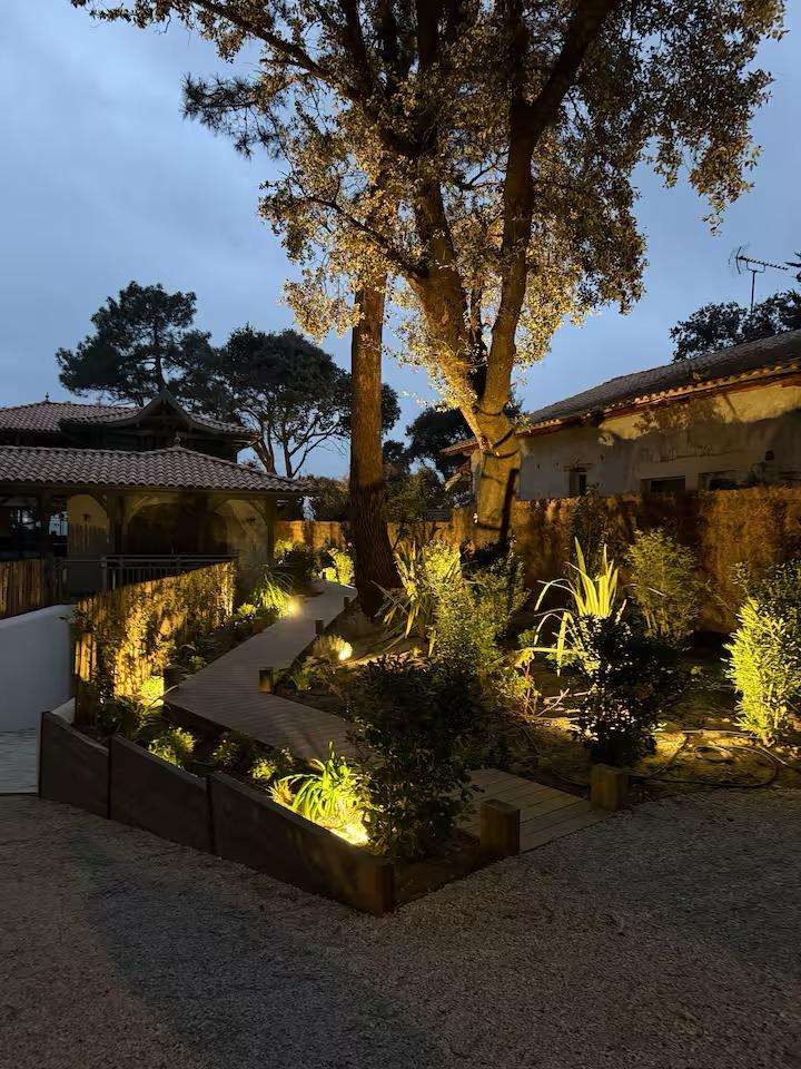 Jardin paysager de nuit avec chemin en bois éclairé, arbres illuminés et maison traditionnelle au crépuscule.