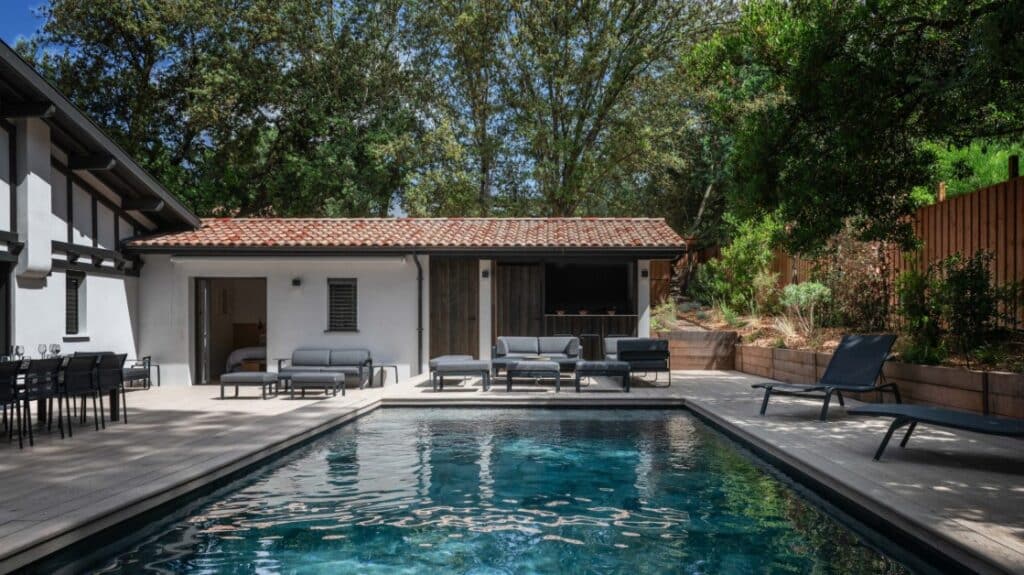 Piscine moderne sur terrasse en bois avec mobilier de jardin gris et pool house blanc entouré de grands arbres.